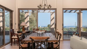 Zona pranzo con un grande tavolo in legno e sedie, un lampadario e vista sul mare tramite ampie vetrate.