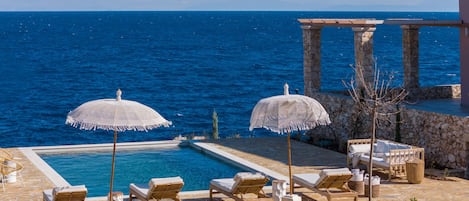 A poolside area with two white umbrellas, lounge chairs, and a stone structure with columns.