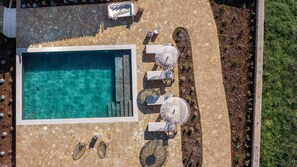 Vista aerea di una zona piscina con terrazza in pietra, lettini e un tavolino con ombrelloni.