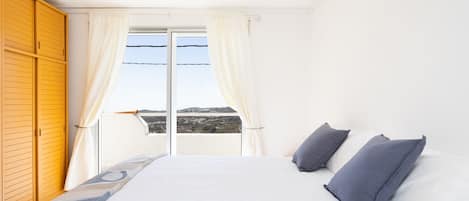 Un dormitorio con una cama grande, ropa de cama blanca, dos almohadas, vistas a través de un ventanal corredero y un armario de madera.