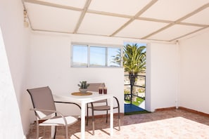 Un pequeño patio amurallado con una mesa y sillas, una ventana y una puerta que da a un jardín con palmeras.