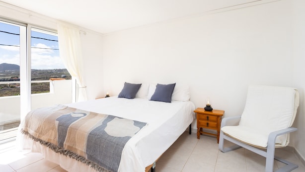 A bedroom with a large bed, a nightstand, a chair, and a view of the landscape. - Oasis Tenerife Apartamento (Granadilla de Abona)
