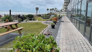 A balcony with a view of the ocean, a grassy area with potted plants, and a wooden picnic table. - Double King Lockout Studios - Steps to the Beach 🏖️ Tiki 🍹 Pool 🌊 (Daytona Beach)