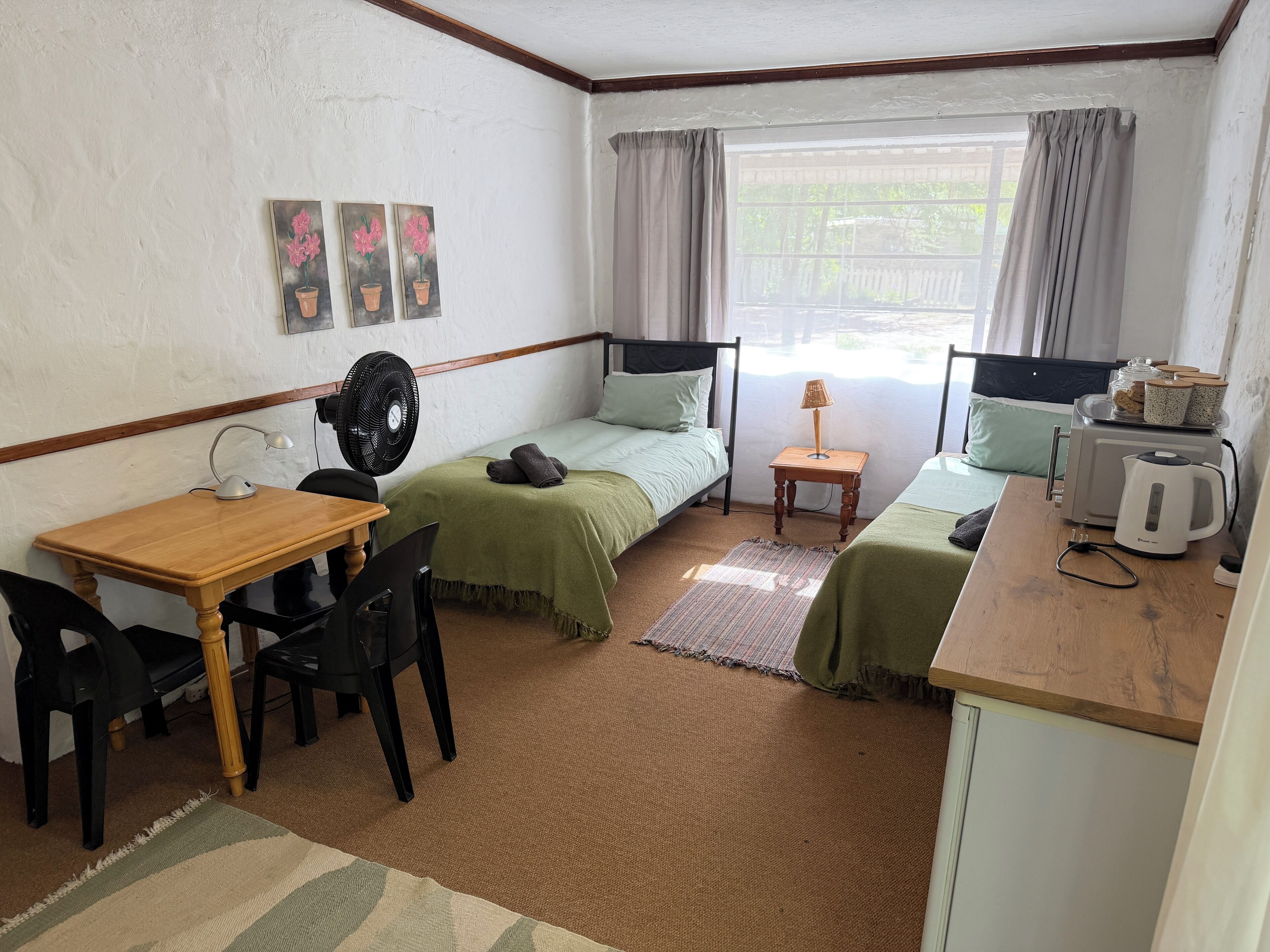 Habitación pequeña con dos camas individuales, una mesita con una silla, un escritorio, un ventilador, una lámpara, un hervidor de agua y una ventana con cortinas.
