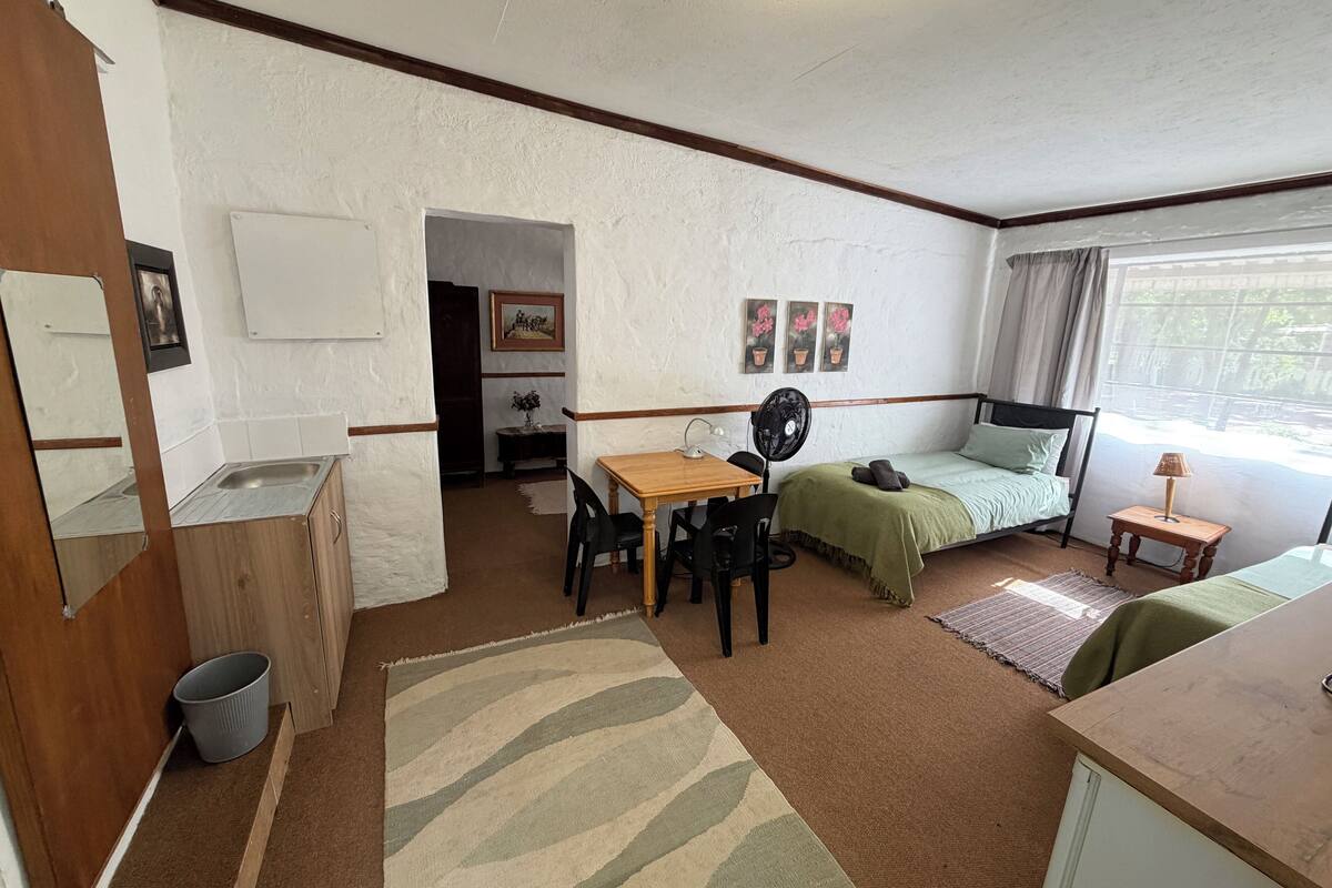 Une pièce de taille modeste comprenant un lit, une petite table avec des chaises, un bureau, un ventilateur, une fenêtre avec des rideaux et un coin cuisine.