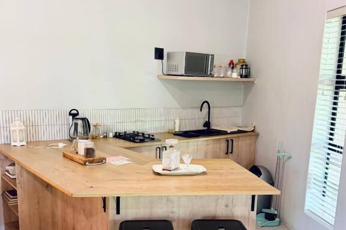 Une cuisine compacte avec un îlot central en bois, un four à micro-ondes et un évier. L’îlot est agrémenté de deux tabourets, et une fenêtre avec des stores est également présente.