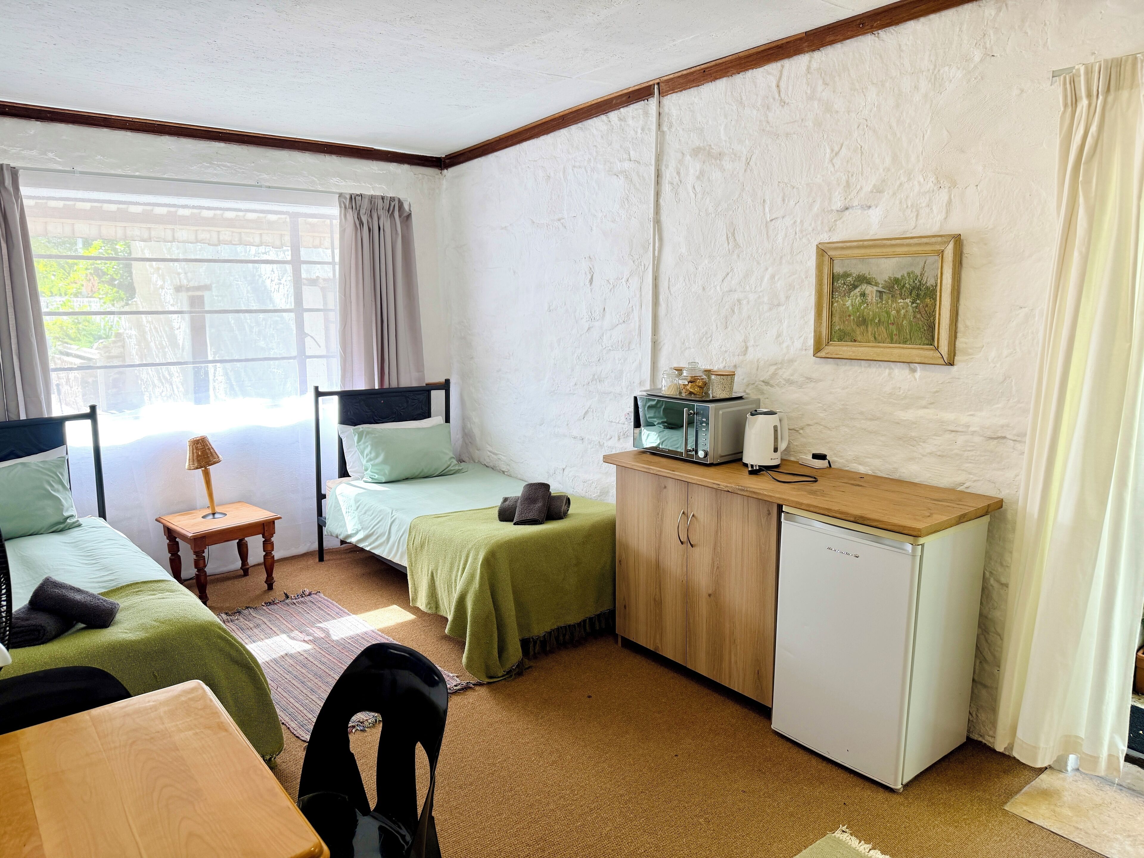 Una habitación pequeña y bien iluminada con dos camas, un kitchenette, un escritorio de madera y un refrigerador pequeño.