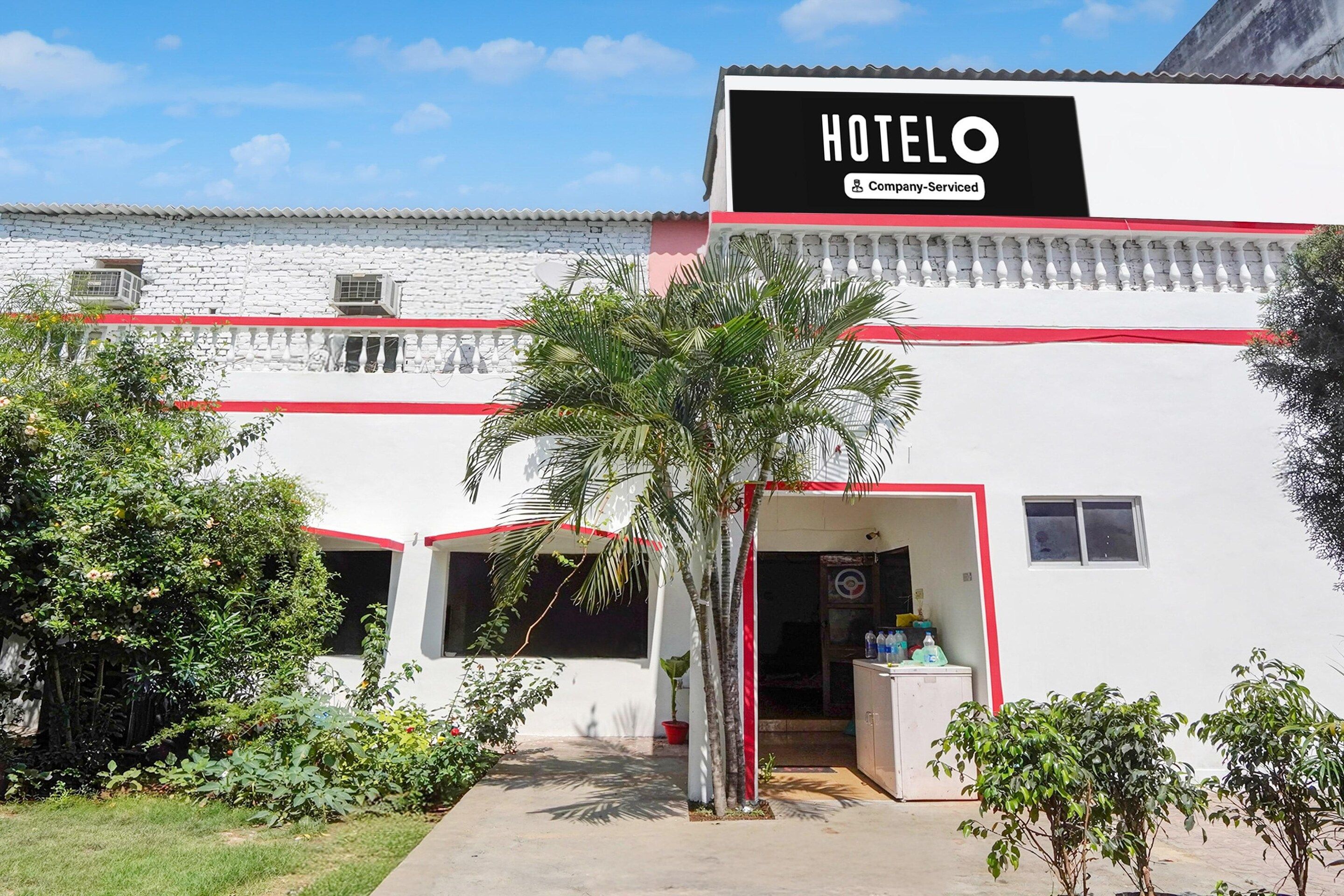 A hotel named 'HOTELO' with a company-serviced sign, a white exterior, and a red trim. There are potted plants and a clear sky.