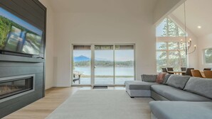 Una sala de estar moderna con chimenea, un sofá gris, un comedor y un amplio ventanal corredera con vista al lago.