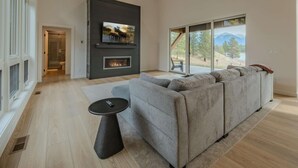 Una sala de estar moderna con chimenea, un sofá seccional grande de color gris, un televisor de pantalla plana montado en la pared y un ventanal con vistas a un paisaje montañoso.