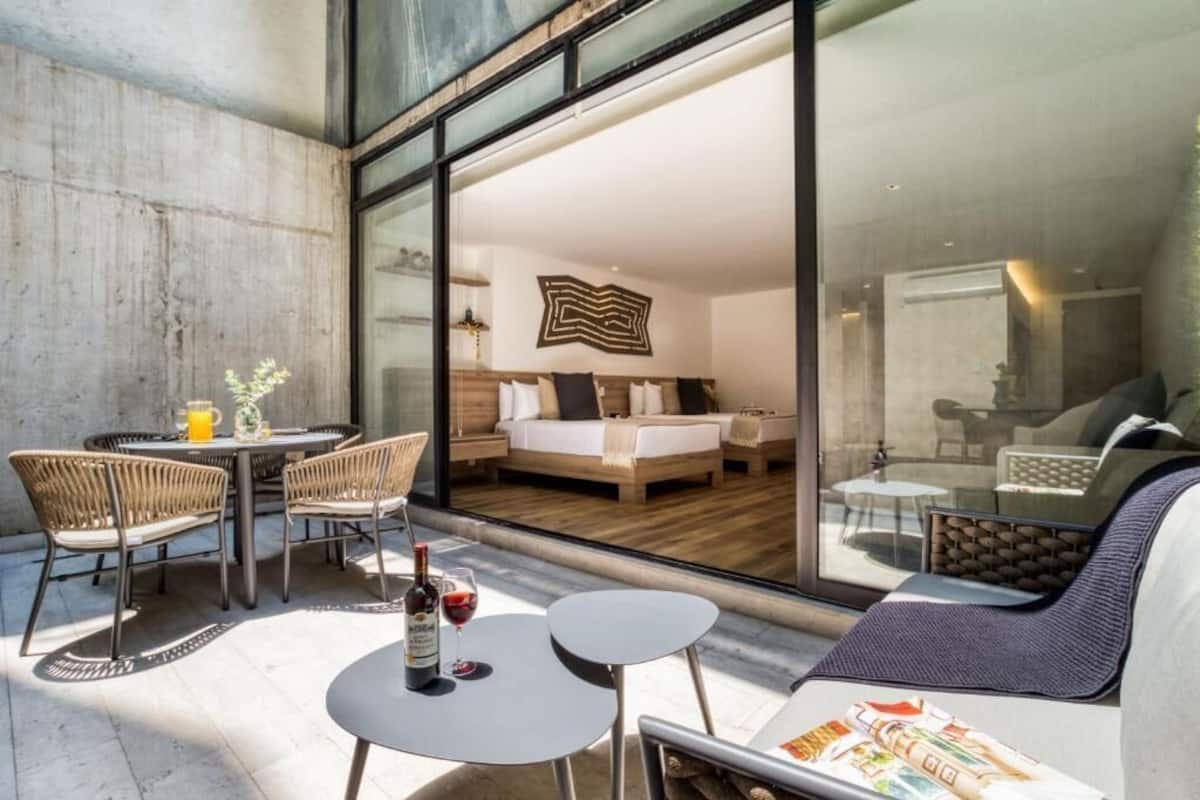 A modern hotel room with a large glass door leading to a balcony, furnished with wicker chairs, a small round table, and a sofa.