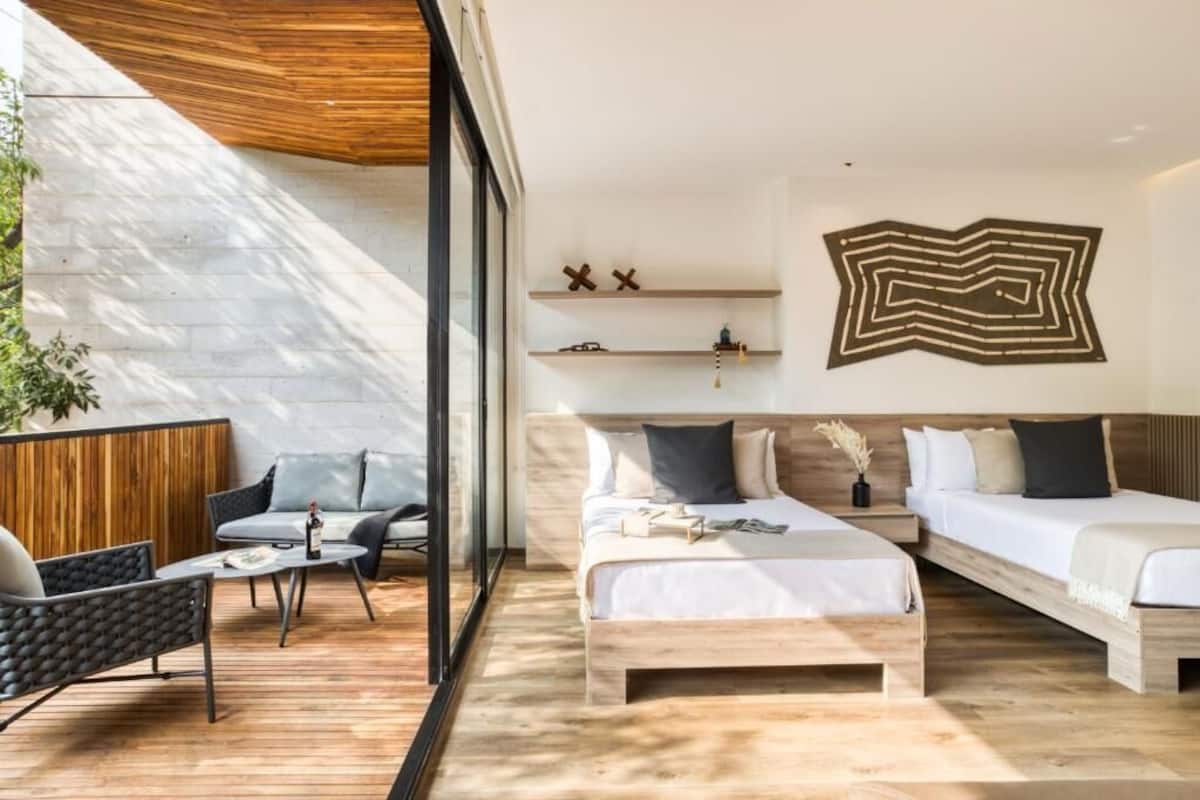 A modern hotel room with a large bed, a smaller bed, a sofa, and a coffee table. There are two shelves with decorative items and a unique wall art piece.