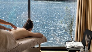 Ein Spa-Behandlungsraum mit großem Fenster und Blick auf ein Gewässer, einer Massageliege und einem kleinen Tisch mit Flasche und Schilferdiffusor.