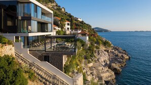 Ett modernt hus vid kusten med en balkong i glas som vetter mot havet, en trappa i sten och utsikt över kustlinjen.