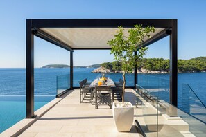 Una piscina a sfioro con vista libera sul mare, un'area pranzo con tavolo apparecchiato per due e una pianta in vaso.
