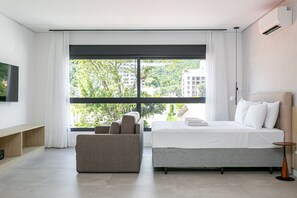 A modern hotel room with a large bed, a sofa, a TV, and a window with a city view.