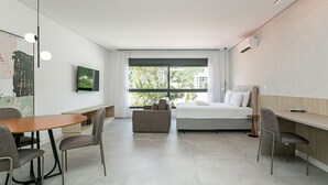A modern hotel room with a bed, a desk, a dining table with chairs, and a large window offering a view of the outdoors. - Near the shopping center | Tech hub | Pool #SG05 (Florianópolis)