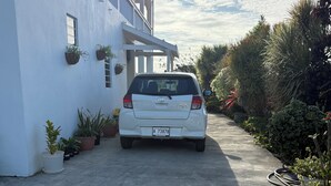 En vit bil står parkerad på en uppfart. Till vänster syns en vit byggnad och till höger finns olika sorters krukväxter.