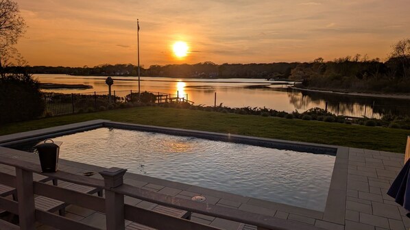 Une piscine avec vue sur un coucher de soleil se reflétant sur un lac, entourée d’une pelouse et d’une clôture.