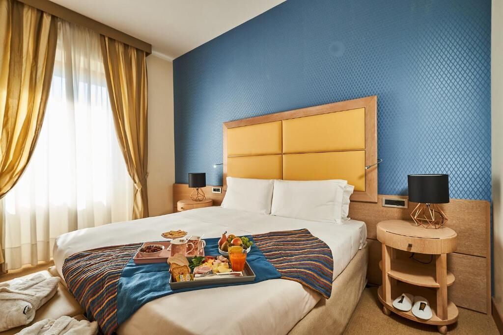 Une chambre d’hôtel avec un grand lit, une table de chevet avec une lampe et une petite table en bois où sont posées des chaussures.