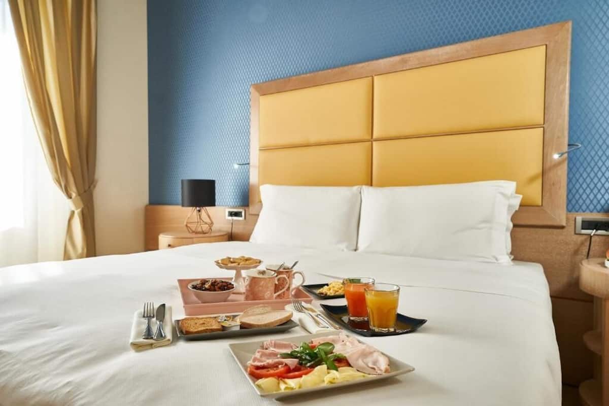 A hotel room with a neatly made bed, breakfast tray with eggs, toast, and juice, and a bedside table with a lamp.