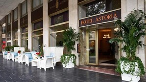 A hotel entrance with a sign reading "HOTEL ASTORIA," outdoor seating with white chairs and blue cushions, and large potted plants. - Hotel Astoria (Biella)