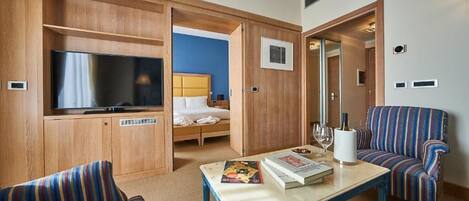 A hotel room with a television, a bed, a sofa, and a table with a book and wine glasses.