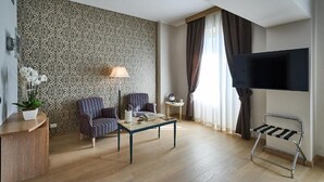 A modern living room with a patterned wall, two striped armchairs, a coffee table, a TV, and a small side table with a lamp. - Hotel Astoria (Biella)