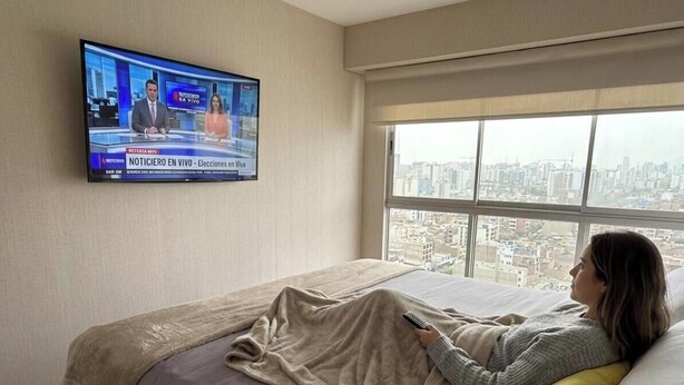 Hotelkamer met een bed, een aan de muur bevestigde televisie waarop een nieuwskanaal draait, en een groot raam met uitzicht op de stad.