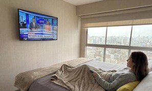 Quarto de hotel com cama, televisão na parede exibindo um canal de notícias e uma janela ampla com vista para a cidade.