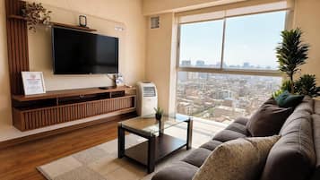 A modern living room with a large flat-screen TV, a sofa, a coffee table, and a view of a cityscape through a large window.