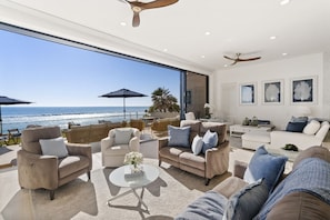 A modern living room with a view of the ocean, furnished with sofas and armchairs, and a ceiling fan. - Luxury on the Beach, Brand New Listing! (Oceanside)