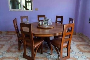 A round wooden dining table with six chairs, a central tray with glasses, and a window above.