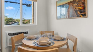 A dining area with a round table set for four, a window with a view of power lines and buildings, and a painting on the wall.