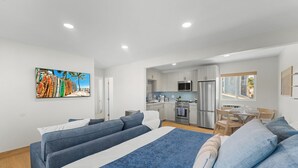 A modern hotel room with a large bed, a kitchenette, a dining area, and a view of the beach.