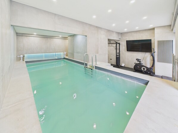 A modern indoor swimming pool with clear water, a changing room with a shower, and a stationary exercise bike.