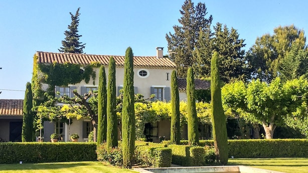 Un edificio grande de dos pisos con tejado de baldosas, rodeado de setos cuidados y árboles altos.