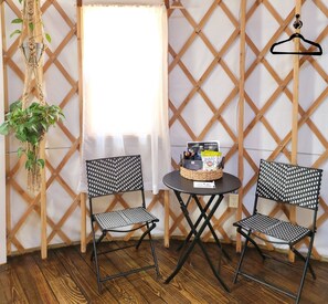 Two black metal chairs with a woven seat and backrest, a small round table with a basket of snacks, and a hanging macrame plant.