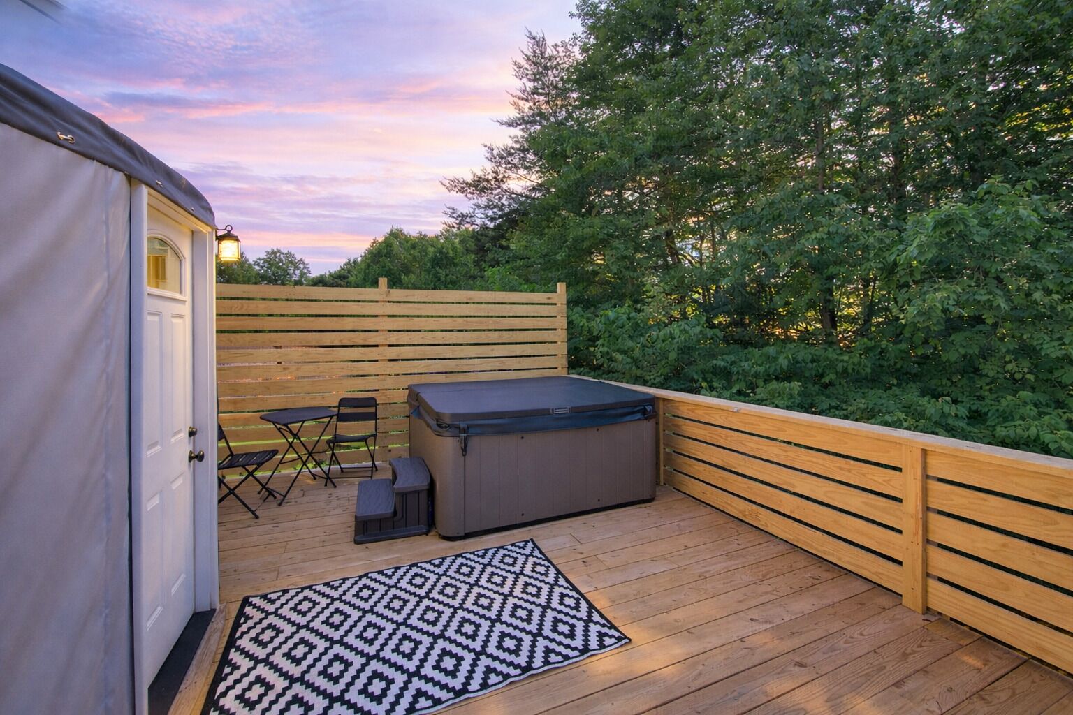 A deck with a hot tub, a table, and chairs, surrounded by a wooden fence and greenery.