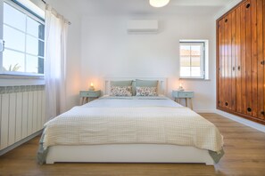 A bedroom with a bed, two bedside tables, a window, a wall-mounted air conditioner, and a wooden wardrobe. - Villa Dos Amores - Heated swimming pool (Azeitão)