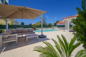 A poolside area with a beige sofa, an umbrella, and a pool. - Villa Dos Amores - Heated swimming pool (Azeitão)