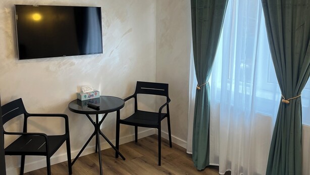 Une chambre d’hôtel équipée d’un téléviseur à écran plat fixé au mur, d’une petite table ronde avec deux chaises noires, d’un lit avec une couette blanche et d’une fenêtre ornée de rideaux verts.