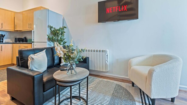 A modern living room with a sofa, a small table, a white armchair, a TV displaying "NETFLIX", and a kitchen in the background. - City Haven Liverpool | 3BR w/ Parking (Merseyside)