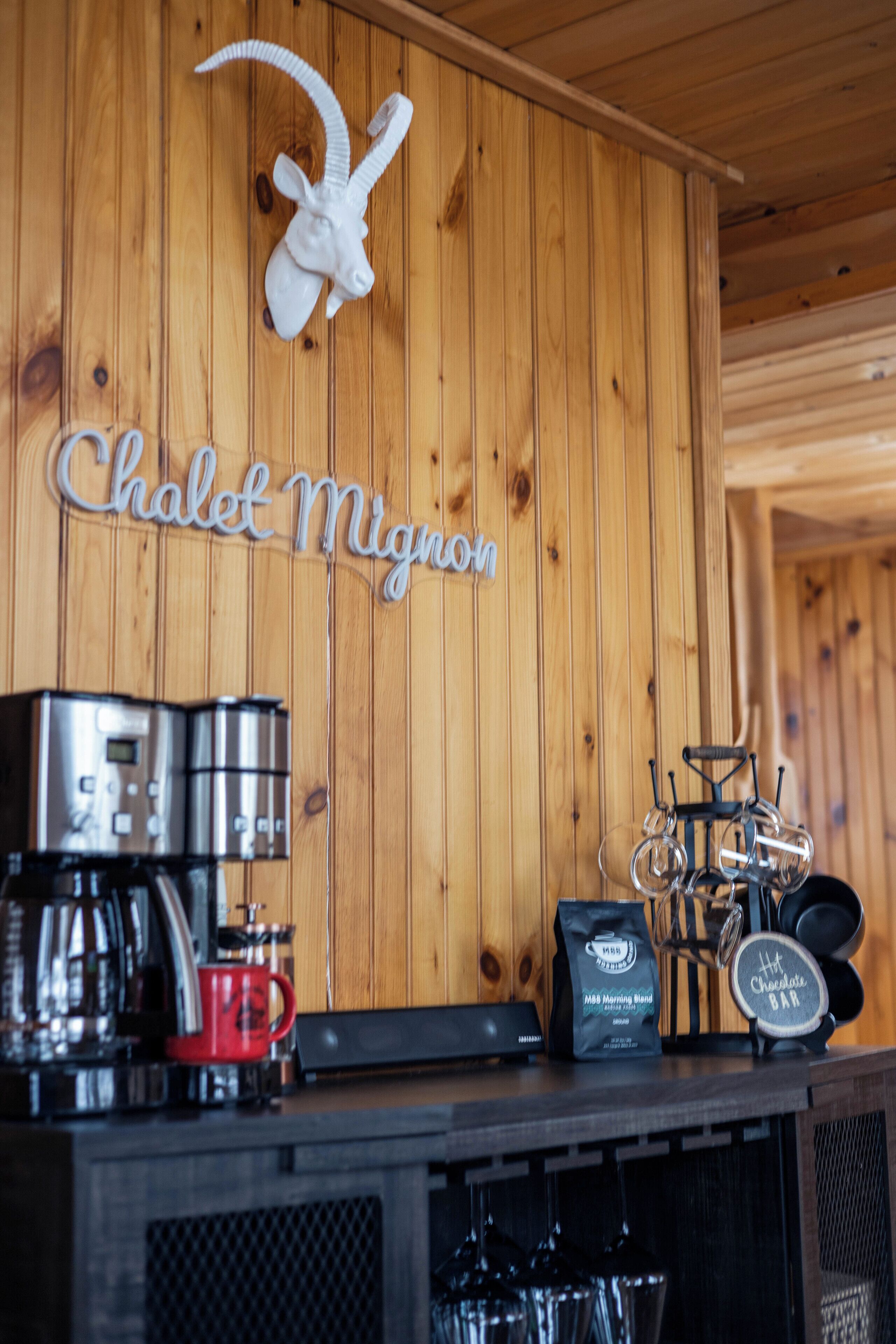 Een houten interieur met een opgehangen dierenhoofd, een koffieapparaat, een rode mok, een tas, en een bordje met de tekst 'Chalet Migron'.