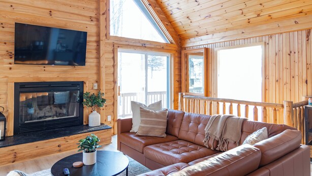 Un interior de cabaña acogedor con un sofá de cuero, una chimenea y ventanas amplias.