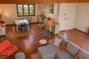 Ein großzügiger Wohnbereich mit grauem Sofa, einem roten Sessel, einem Esstisch mit Stühlen, einer Küche mit Spüle und Schränken sowie einem Fenster mit Blick auf Grünflächen.