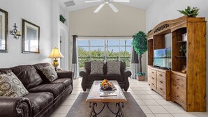 A spacious living room with a high ceiling, a ceiling fan, a large window with a view of greenery, a wooden cabinet with drawers, and a leather sofa with patterned cushions. - Delightful Family Pool Villa in Glenbrook with 4 Bedrooms & 3 Bathrooms (Clermont)