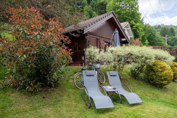 Eine Holzhütte mit geneigtem Dach, umgeben von üppiger Vegetation und zwei Liegestühlen.