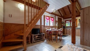 Un escalier en bois mène à un espace repas comprenant une table et des chaises, avec une cuisine en arrière-plan et un téléviseur posé sur un meuble.