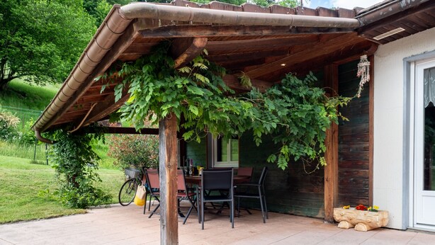 Une terrasse couverte avec une table et des chaises, un vélo et un jardinier en bois orné de fleurs.